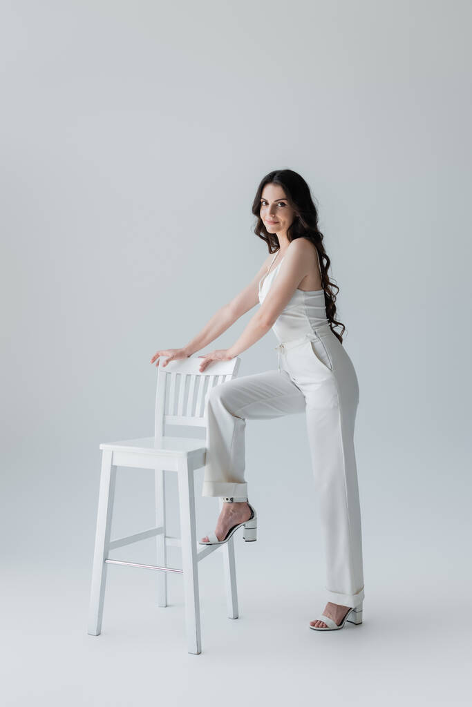 Smiling model in heels and white clothes looking at camera near chair on grey background - Fotoğraf, Görsel