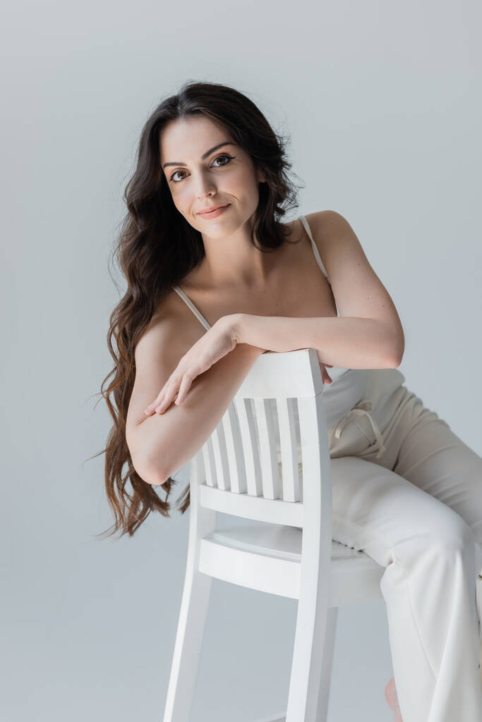 Positive long haired woman sitting on chair isolated on grey  - Foto, afbeelding