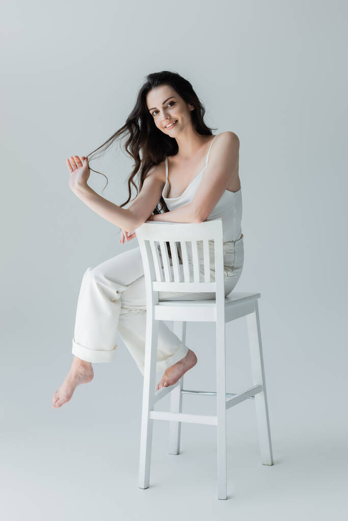Full length of positive barefoot woman sitting on chair on grey background - Foto, immagini