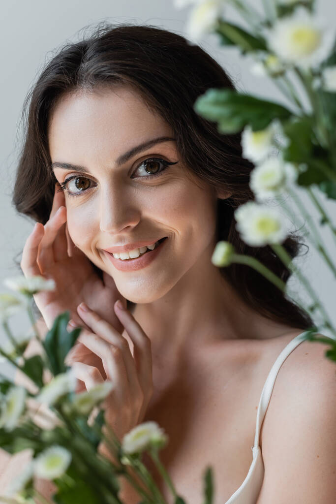 Cheerful brunette model looking at camera near blurred chamomiles isolated on grey  - Photo, image