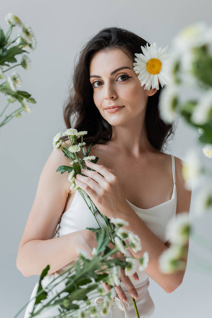 Brunette woman in white top holding chamomiles and looking at camera isolated on grey  - Foto, imagen
