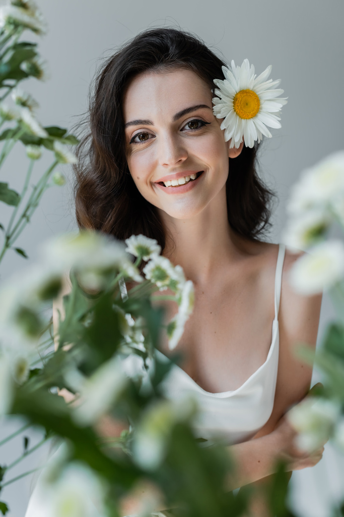 Positive model in white top smiling at camera near blurred chamomiles isolated on grey  - Foto, immagini