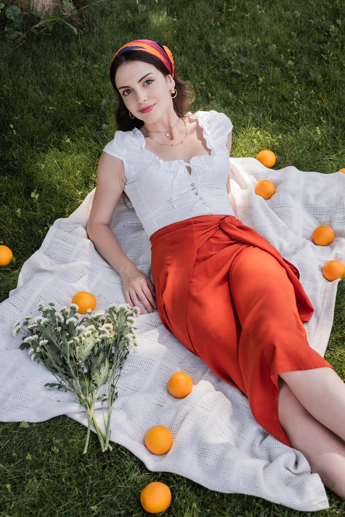 Высокоугольный вид стильной женщины, смотрящей на камеру рядом с цветами и апельсинами на одеяле в парке  - Фото, изображение