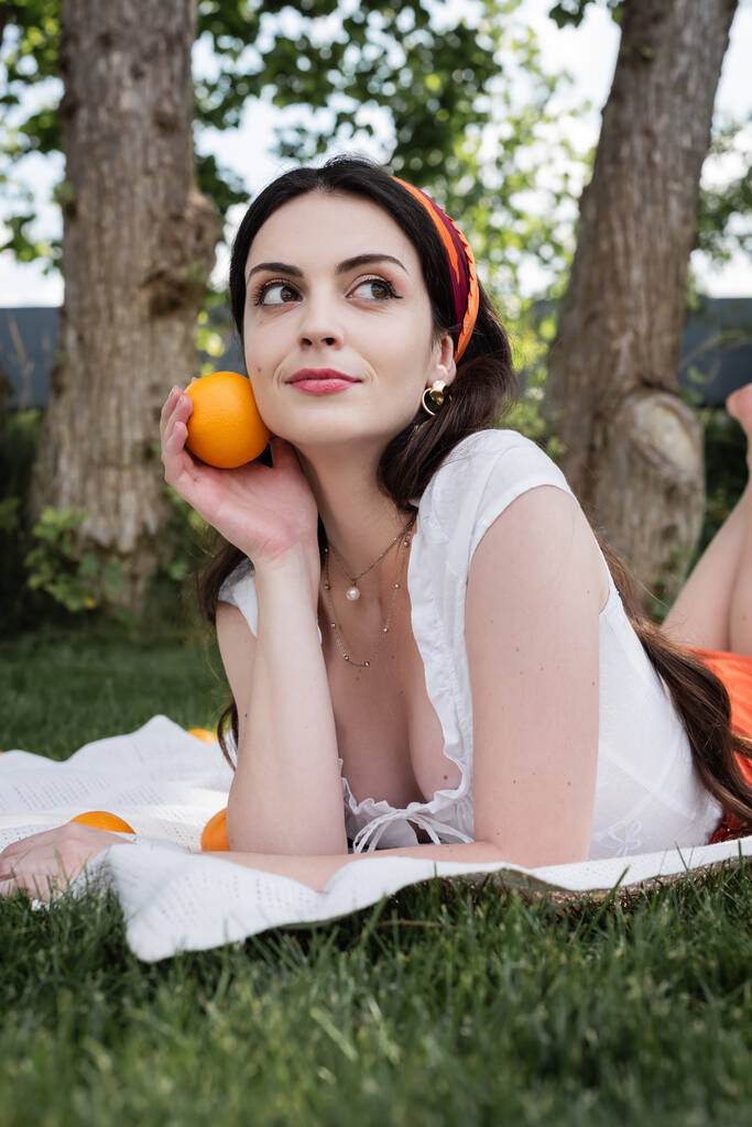 Stylish young woman holding orange on blanket and blurred grass in park  - Foto, immagini