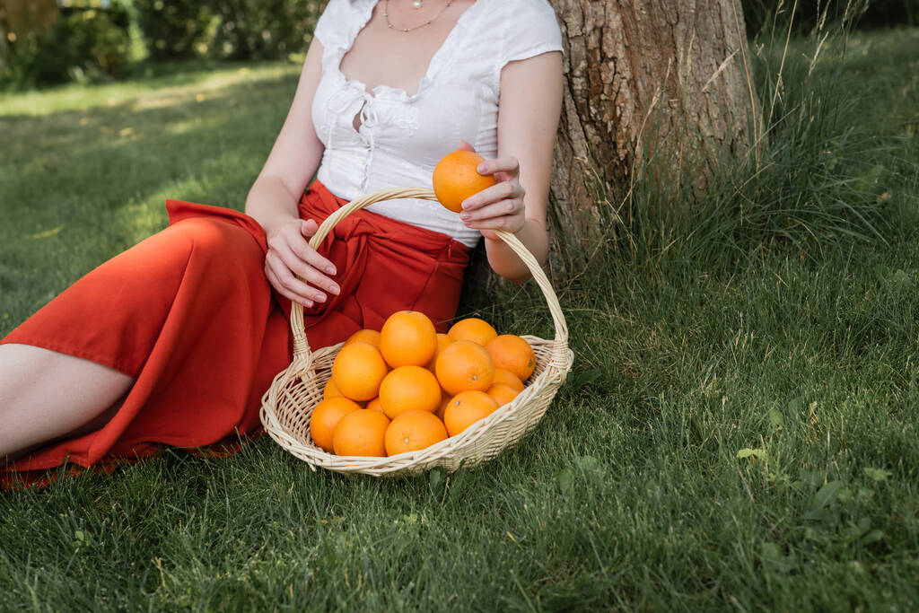 Cropped view of stylish woman holding orange near basket in park  - Foto, imagen