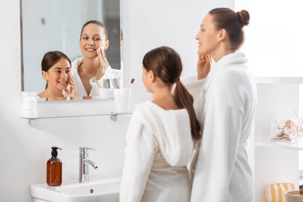 beauty, hygiene, morning and people concept - happy smiling mother and daughter with cotton pads cleaning face skin and looking to mirror at bathroom - 写真・画像