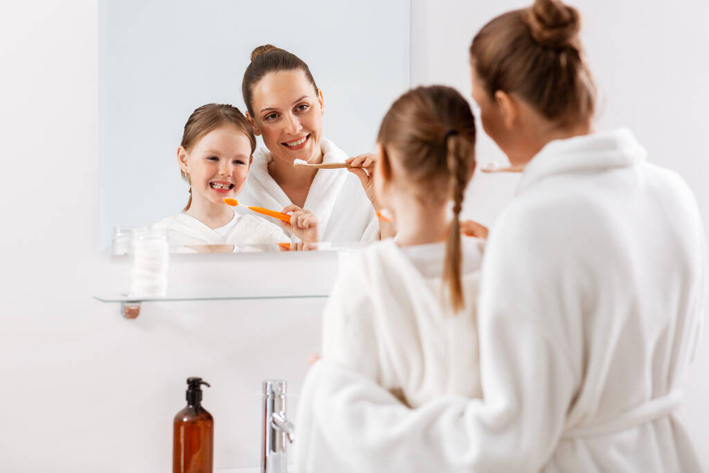 güzellik, hijyen, sabah ve insanlar konsepti - mutlu gülümseyen anne ve küçük kızı diş fırçalarıyla banyoda dişlerini fırçalıyorlar - Fotoğraf, Görsel
