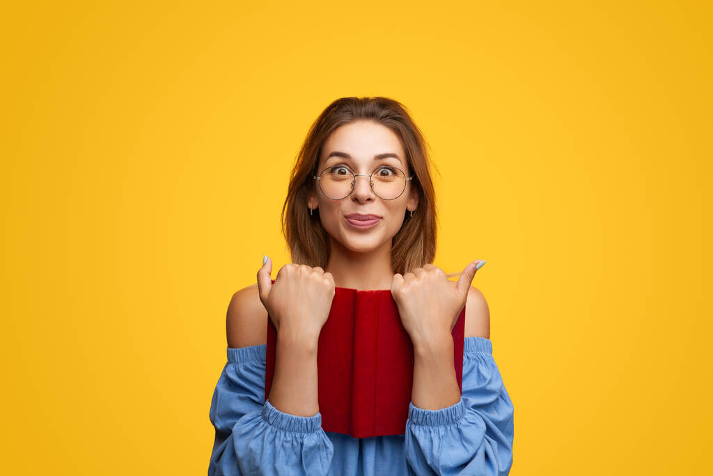 Delighted young smart female student with brown hair in blouse and eyeglasses looking at camera with excitement while holding interesting book against yellow background - Zdjęcie, obraz