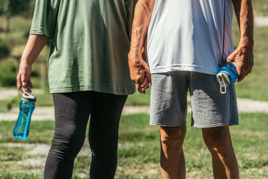 cropped view of retired couple in sportswear with sport bottles holding hands in park - Foto, imagen