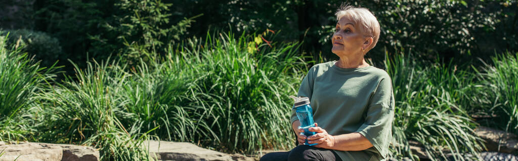 συνταξιούχος γυναίκα στο αθλητικό ντύσιμο κάθεται με αθλητικό μπουκάλι γύρω από πράσινα φυτά, πανό - Φωτογραφία, εικόνα