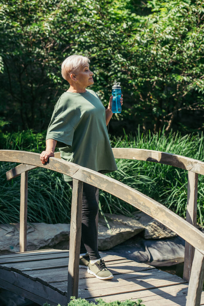 full length of retired woman in sportswear holding sports bottle and standing on bridge - 写真・画像