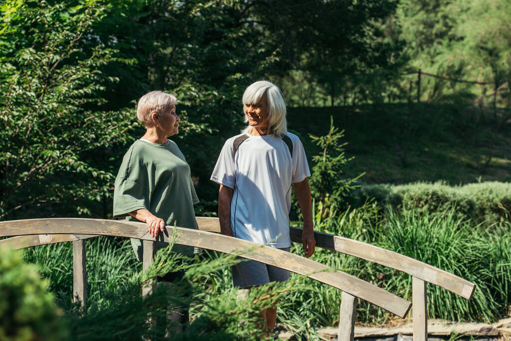 retired senior couple in sportswear smiling while standing on bridge  - 写真・画像