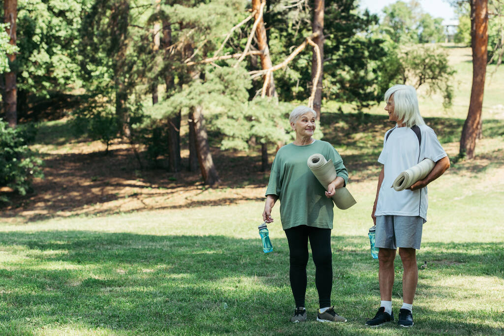 повна довжина весела старша пара в спортивному одязі, що тримає фітнес-мати і спортивні пляшки в парку
  - Фото, зображення