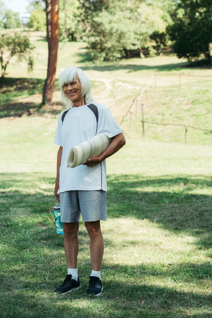 full length of happy senior man in white t-shirt holding sports bottle and fitness mat in park - Zdjęcie, obraz