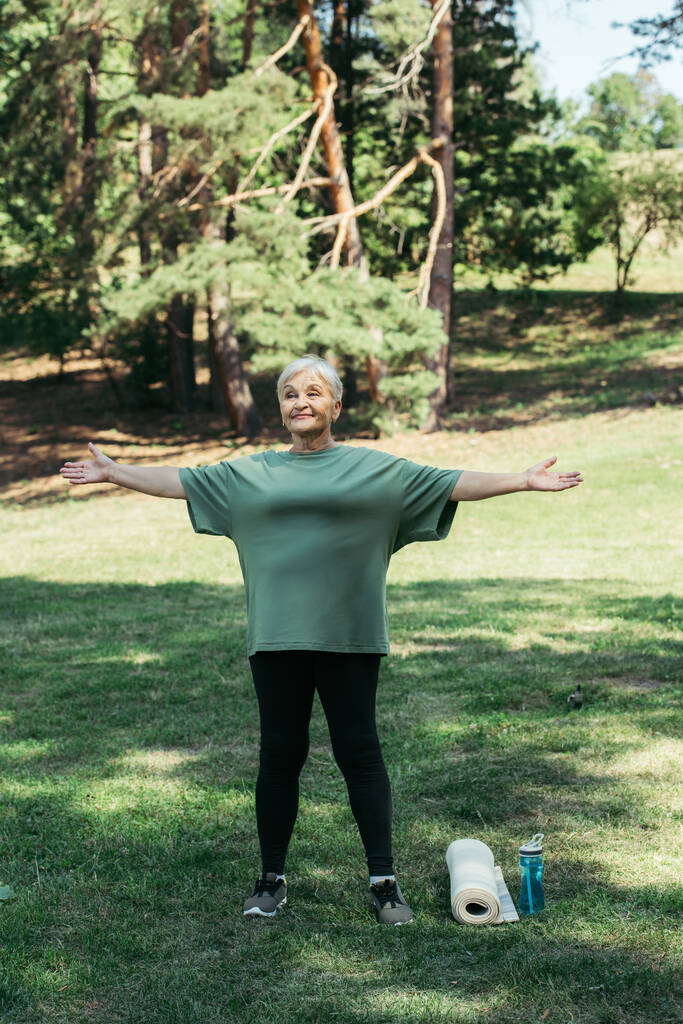 full length of happy senior woman exercising near sports bottle and fitness mat in park - Fotó, kép