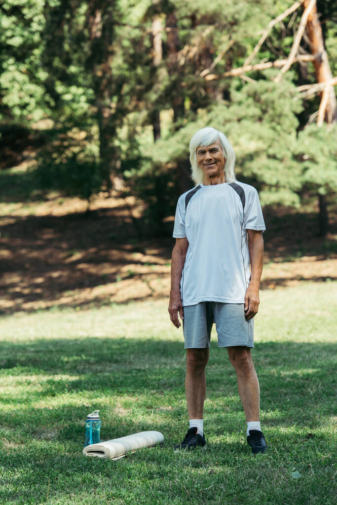 full length of happy senior man in sportswear standing near sports bottle and fitness mat on grass - Fotografie, Obrázek