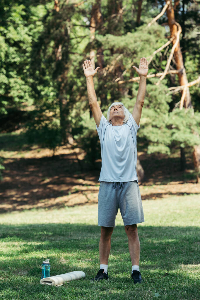 full length of happy senior man in sportswear exercising near sports bottle and fitness mat on grass - Fotó, kép