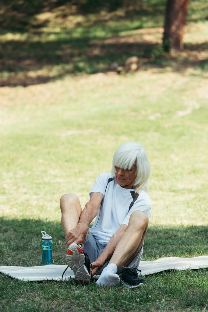 senior man with grey hair taking off sneakers while sitting on fitness mat in park - Photo, Image