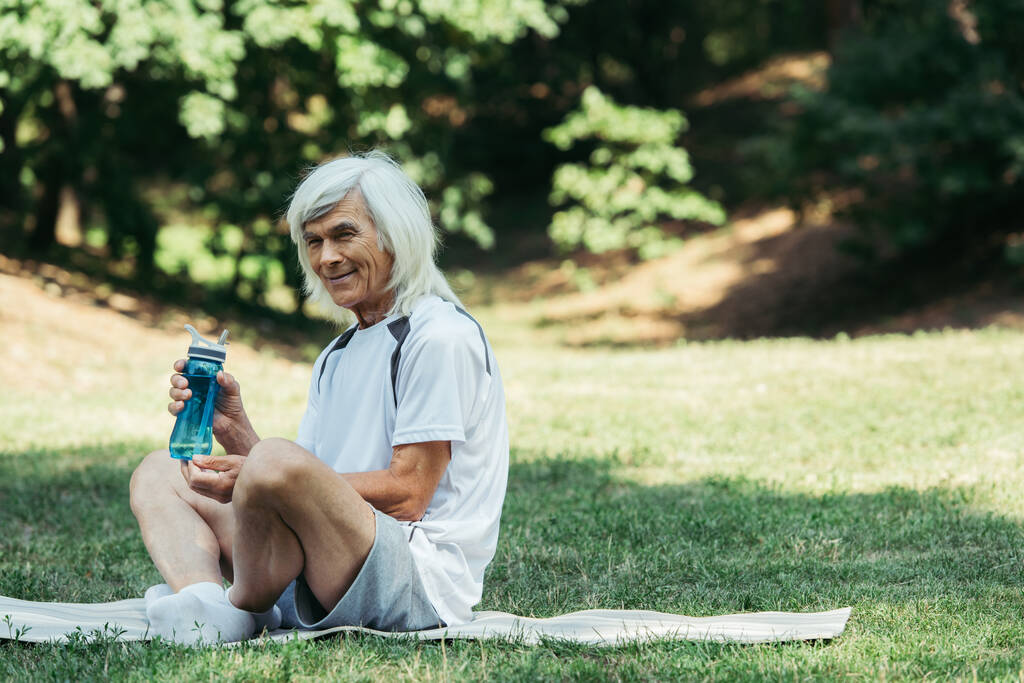 joyful senior man with grey hair holding sports bottle while sitting on fitness mat in green park - Foto, afbeelding