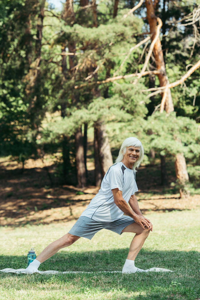 full length of senior man with grey hair smiling and doing lunges on fitness mat in park - Foto, immagini