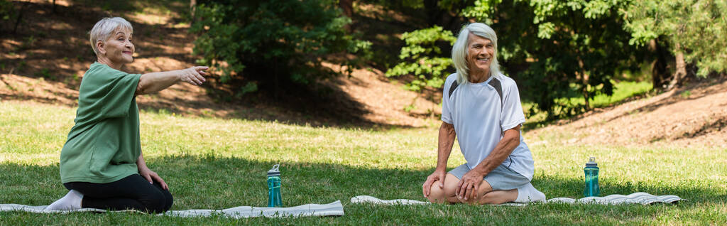 full length of senior woman pointing with finger near happy husband on fitness mat in green park, banner - Fotó, kép