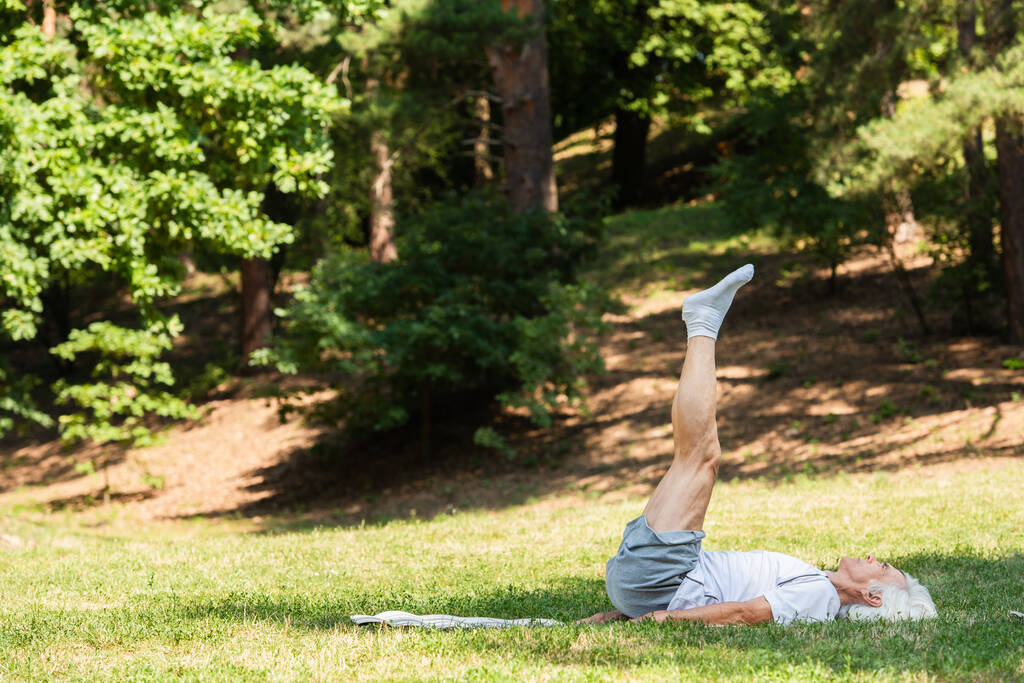 full length of senior man in sportswear doing shoulder stand on fitness mat in green park  - Photo, Image