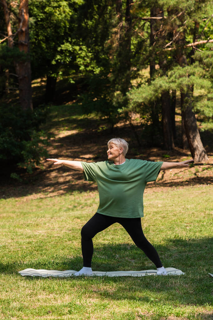 Пожилая женщина в полный рост с седыми волосами улыбается и тренируется на фитнес-коврике в парке - Фото, изображение