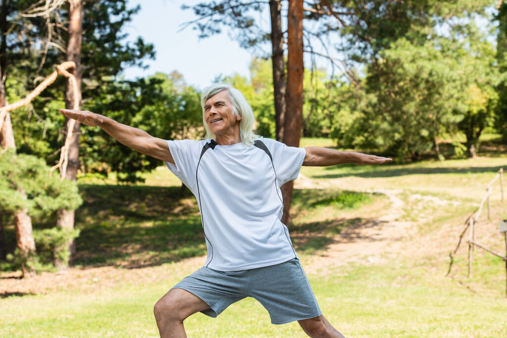 cheerful senior man with grey hair smiling and working out with outstretched hands in park - Photo, image