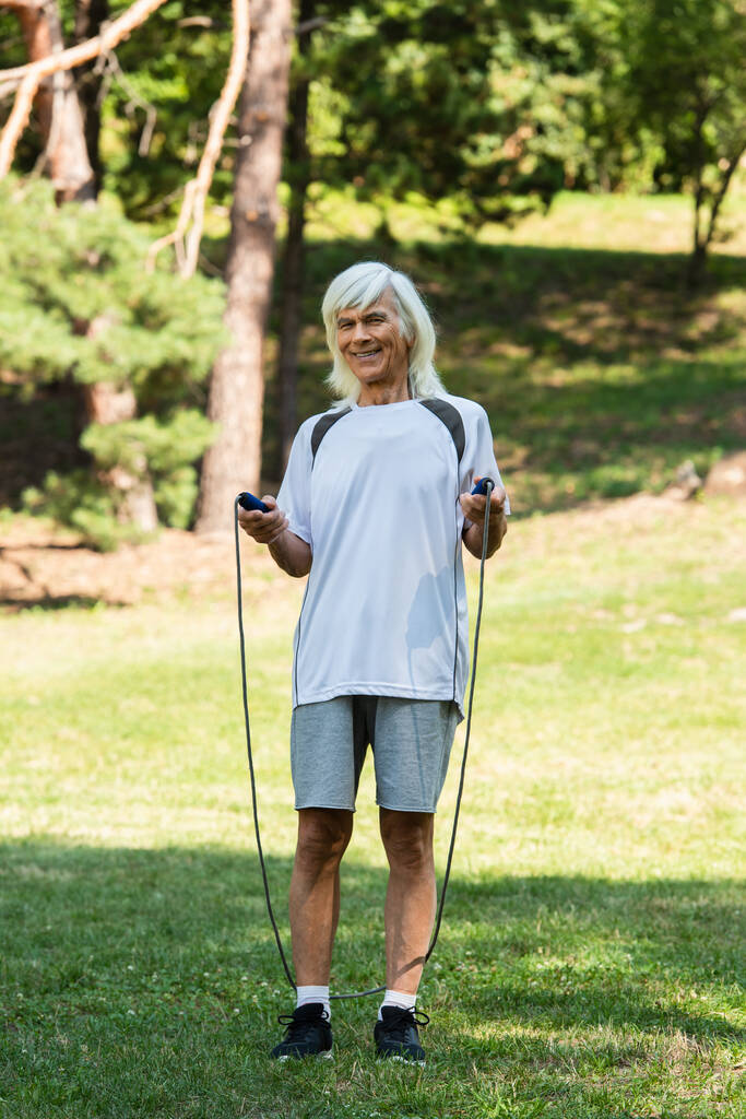 full length of happy senior man with grey hair holding skipping rope in park - Zdjęcie, obraz