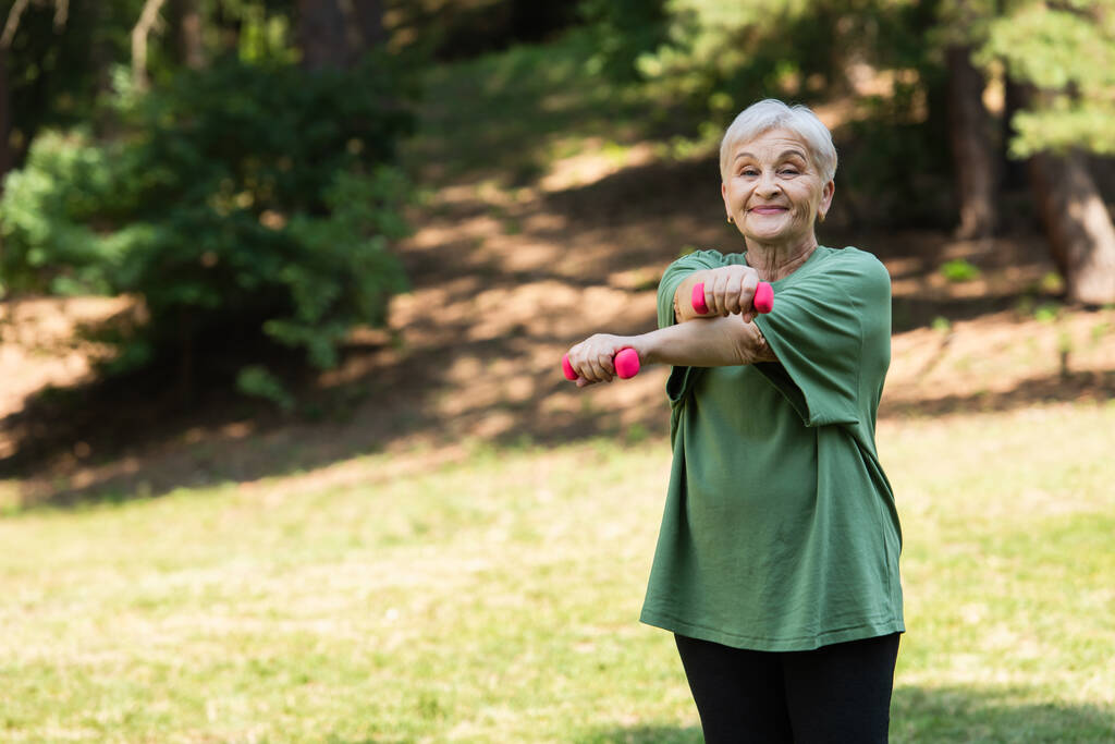 щаслива старша жінка з сірим волоссям посміхається і займається з гантелями в парку
 - Фото, зображення