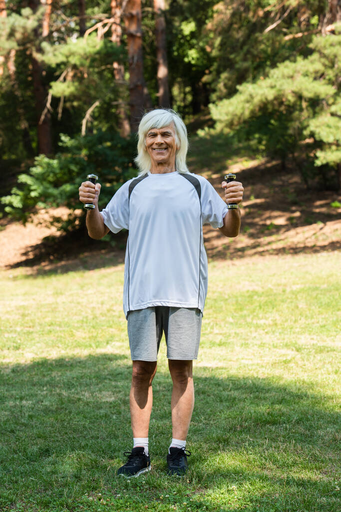 full length of happy senior man in sportswear working out with dumbbells in green park  - Fotografie, Obrázek