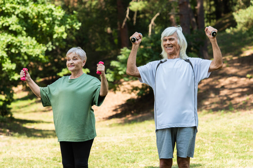 cheerful senior couple in sportswear exercising with dumbbells in green park  - Foto, imagen