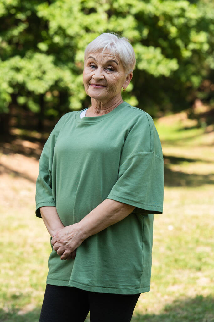 cheerful and retied woman with grey hair standing in green t-shirt in park - Foto, Bild