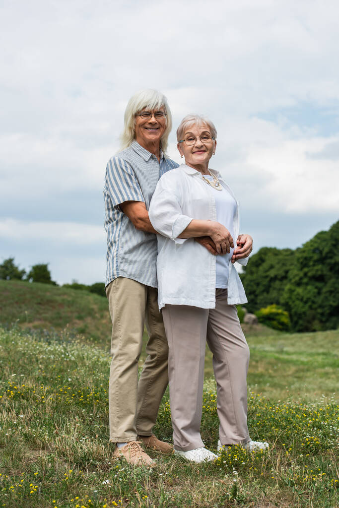joyful senior man in glasses hugging happy wife with grey hair and standing on green hill  - Foto, imagen