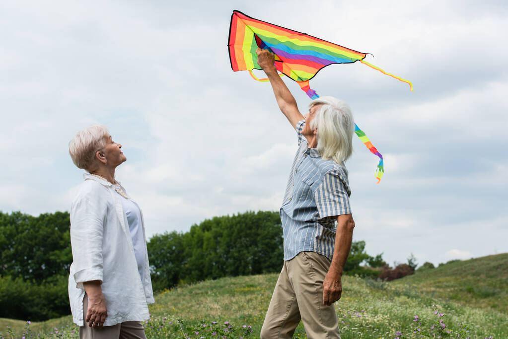 ευτυχισμένο ζευγάρι ηλικιωμένων σε casual ρούχα κοιτάζοντας ιπτάμενο χαρταετό και στέκεται στο πράσινο λόφο  - Φωτογραφία, εικόνα