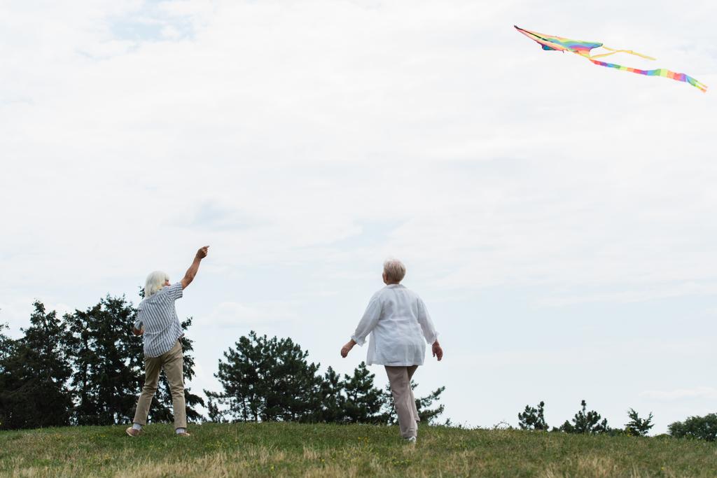 senior woman in casual clothes walking near husband playing with kite on green hill  - Photo, Image