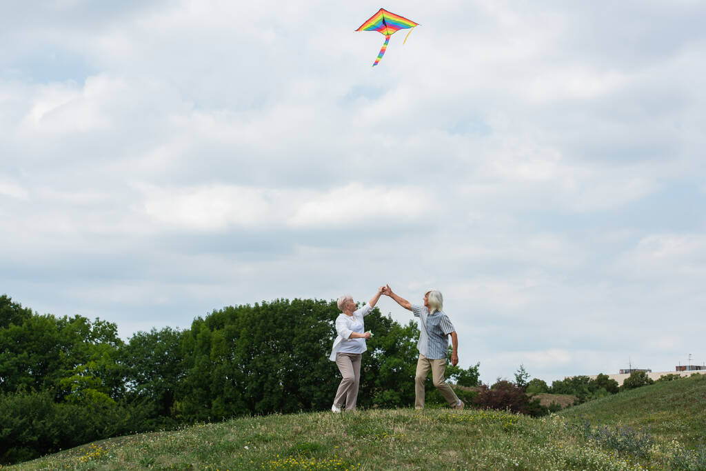 веселая старшая пара в повседневной одежде, держась за руки, играя с воздушным змеем на зеленом холме  - Фото, изображение