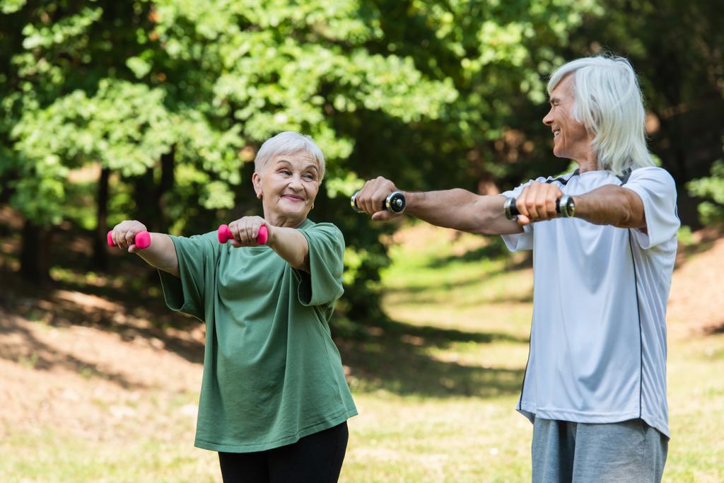 positive retired couple in sportswear exercising with dumbbells in green park  - Zdjęcie, obraz