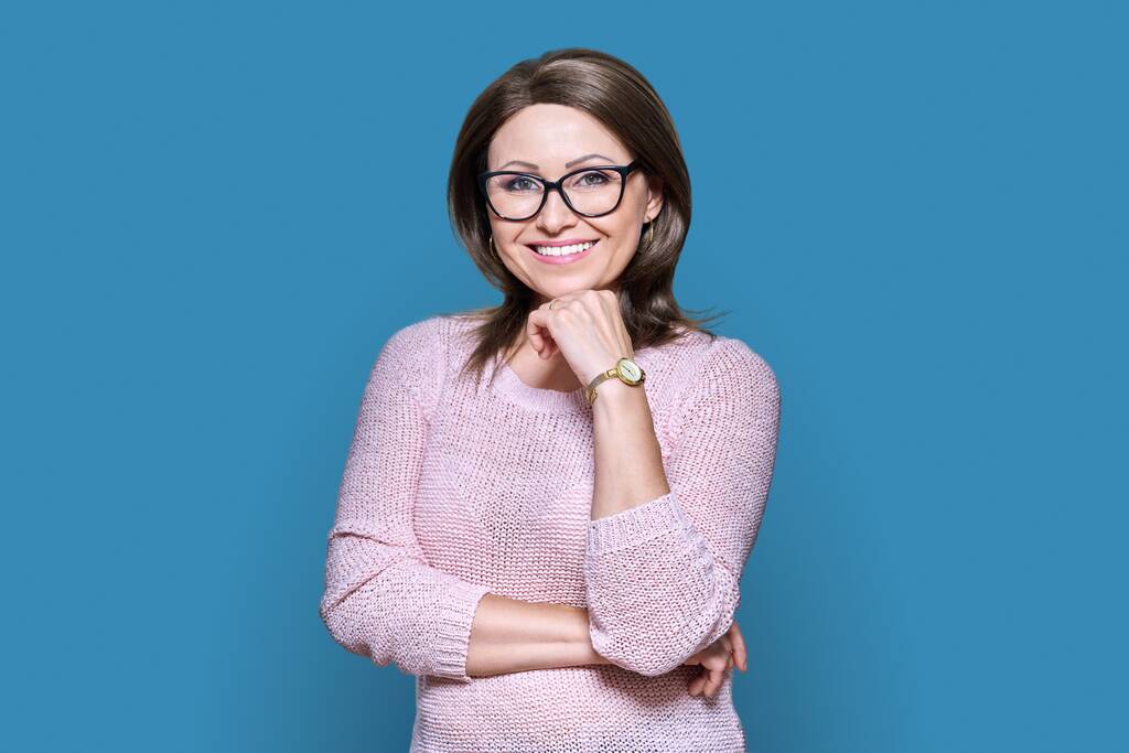 Attractive confident middle aged woman looking at camera on blue color background. Smiling 40s female with glasses, with hand near face. Lifestyle, confidence, calmness, mature people concept - Zdjęcie, obraz