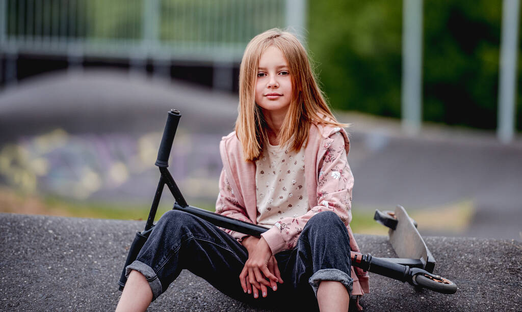 Preteen κορίτσι με σκούτερ κάθεται στο πάρκο της πόλης την άνοιξη και κοιτάζοντας την κάμερα. Όμορφο παιδικό μοντέλο ποζάρουν με το όχημα - Φωτογραφία, εικόνα