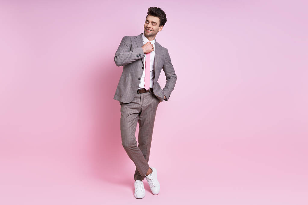 Веселий молодий чоловік у повному костюмі налаштував свою краватку, стоячи на рожевому фоні
 - Фото, зображення