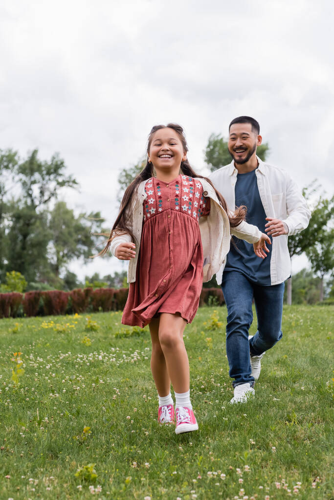 Веселая азиатская девушка бегает рядом с отцом в летнем парке  - Фото, изображение