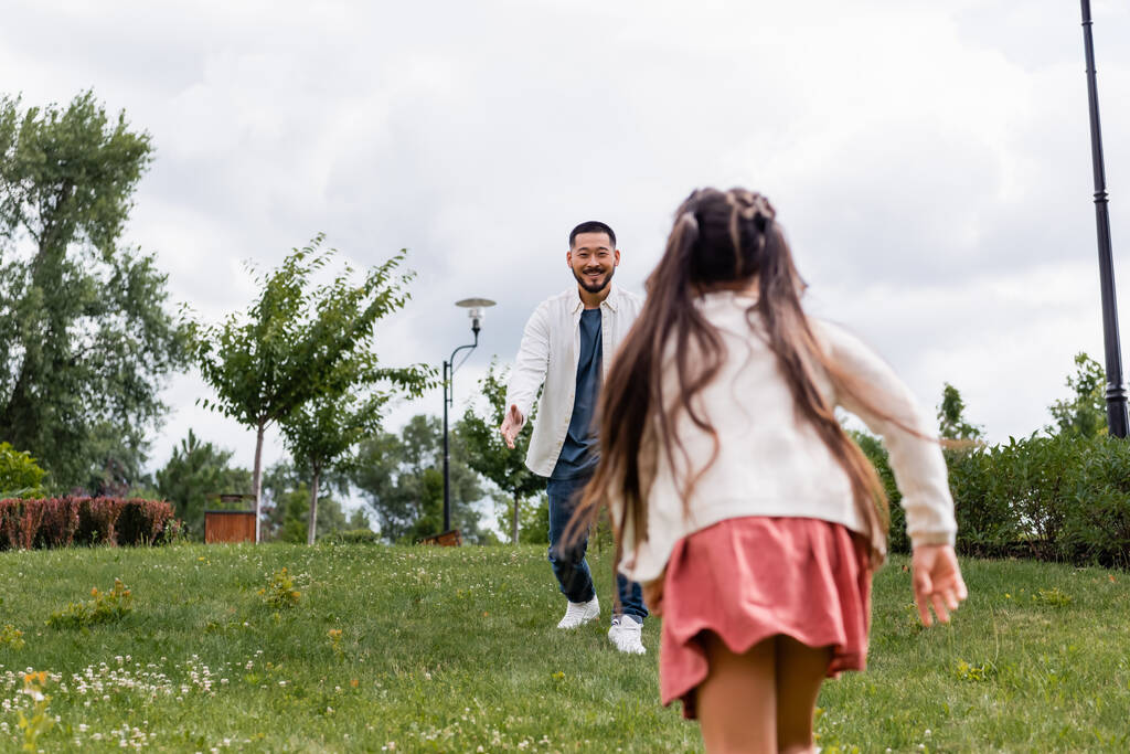 Усміхнений азіатський чоловік дивиться на бігу дочки в літньому парку
  - Фото, зображення