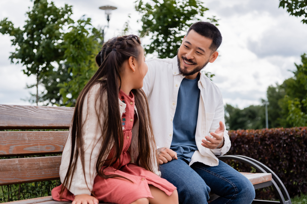 Усміхнений азіатський чоловік розмовляє з дочкою на лавці в парку
  - Фото, зображення