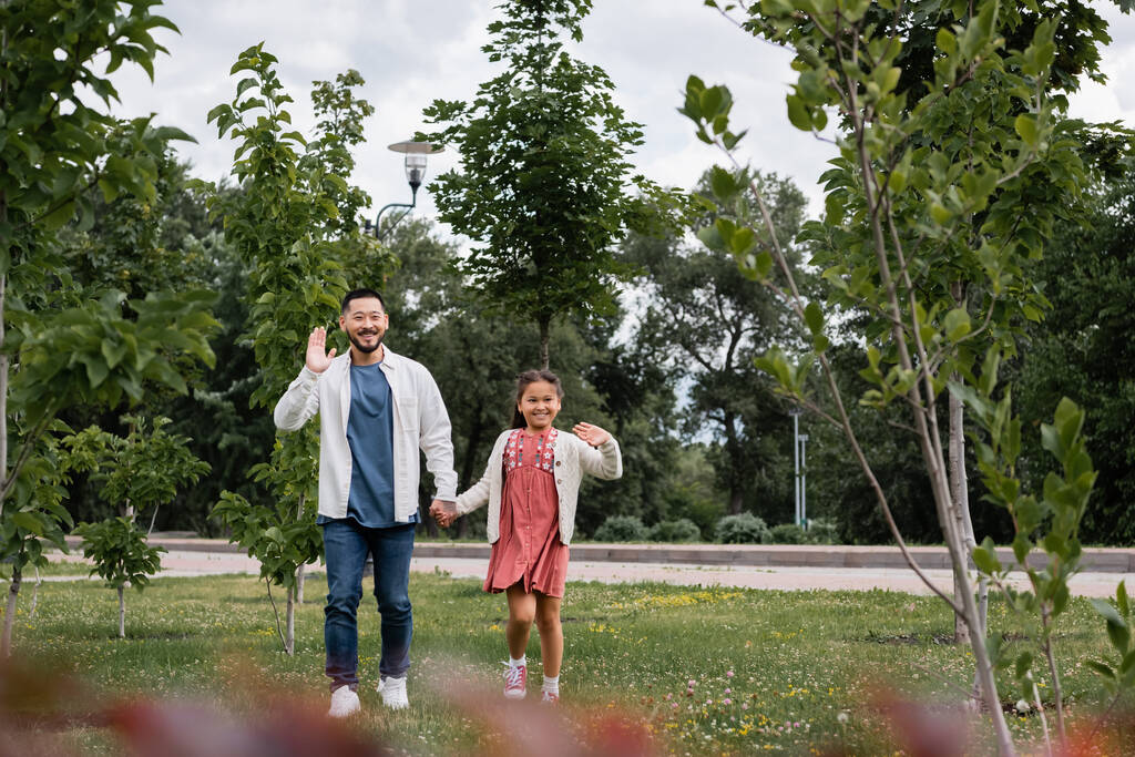 Smiling asian dad and daughter waving hands at camera in park  - Fotó, kép