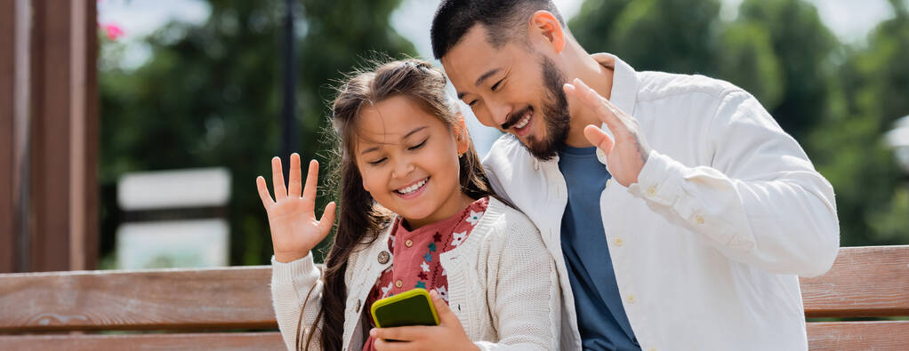 Азіатські батьки і дитина мають відеодзвінок на смартфоні на лавці літнього парку, банер.  - Фото, зображення