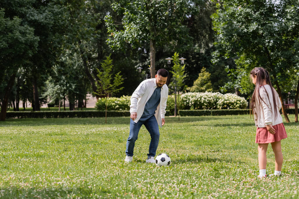 Усміхнений азіатський чоловік грає у футбол з дочкою на газоні в парку
  - Фото, зображення
