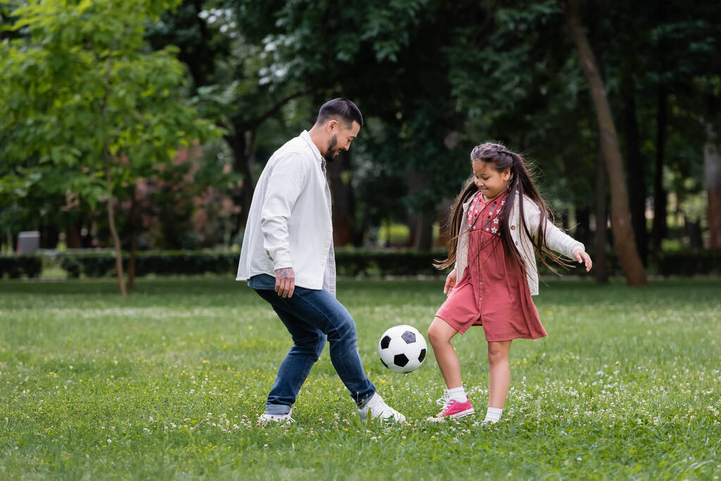 Positive asian kid and parent playing football on meadow in park  - 写真・画像