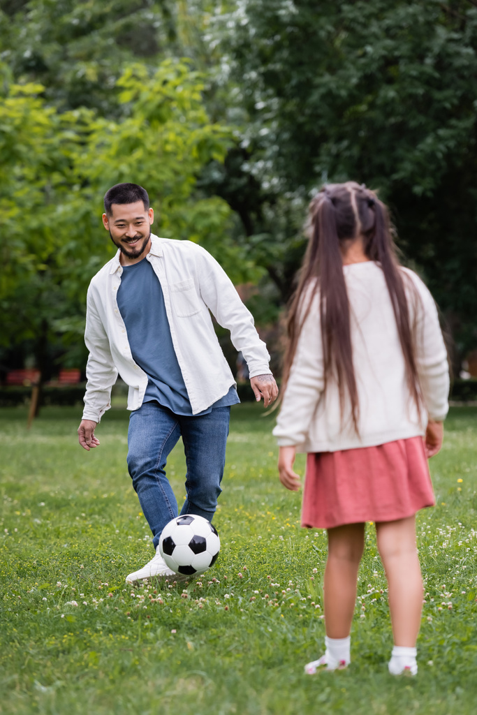 Cheerful asian dad playing football with blurred daughter in park  - Photo, image