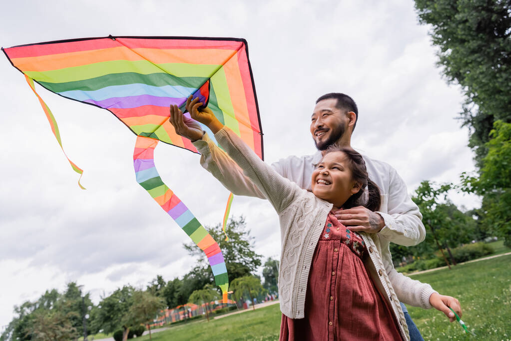 Positive asian dad hugging daughter with flying kite in summer park  - Fotografie, Obrázek